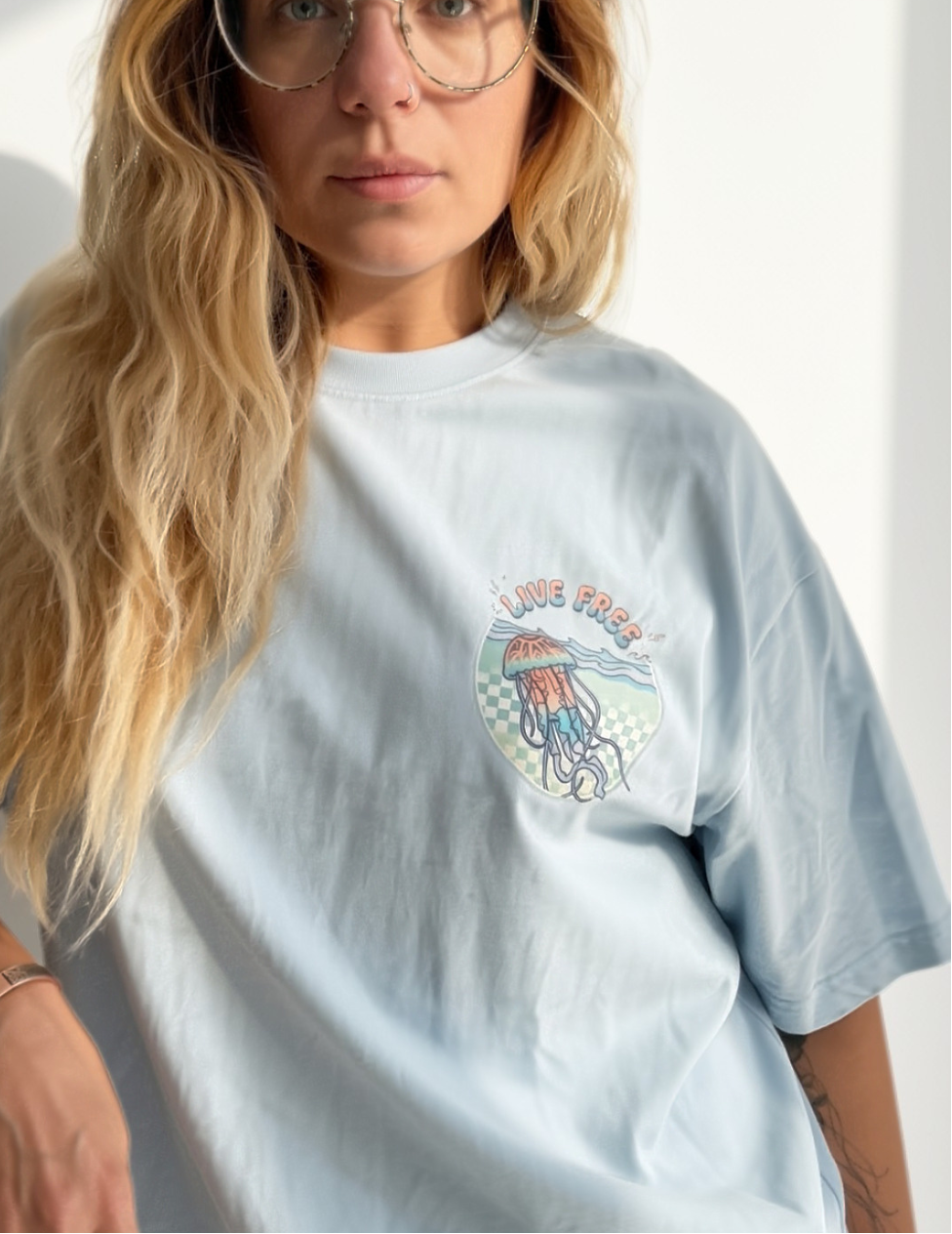 A woman with long blonde hair and glasses wearing a light blue oversized t-shirt with a jellyfish graphic on the chest, set against a bright white background with soft shadows.