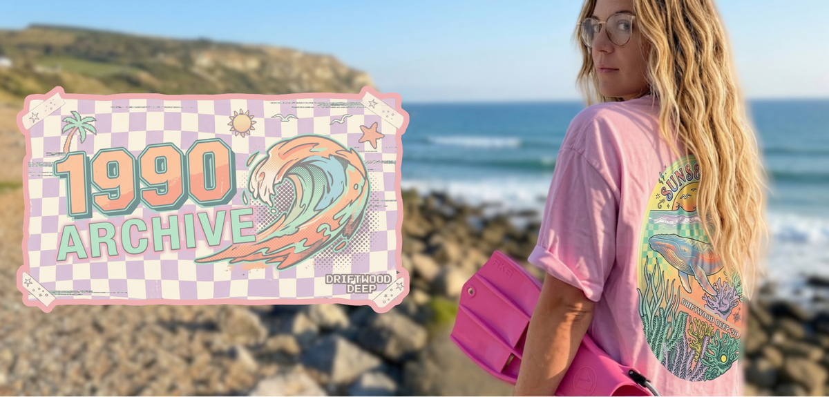 A woman scuba diver stands on a rocky beach wearing a retro pink graphic T-shirt called the Sunseeker Tee with a large multicolour humpback whale design on the back. She is holding pink scuba diving fins, with the ocean and Folkestone cliffs in the background.