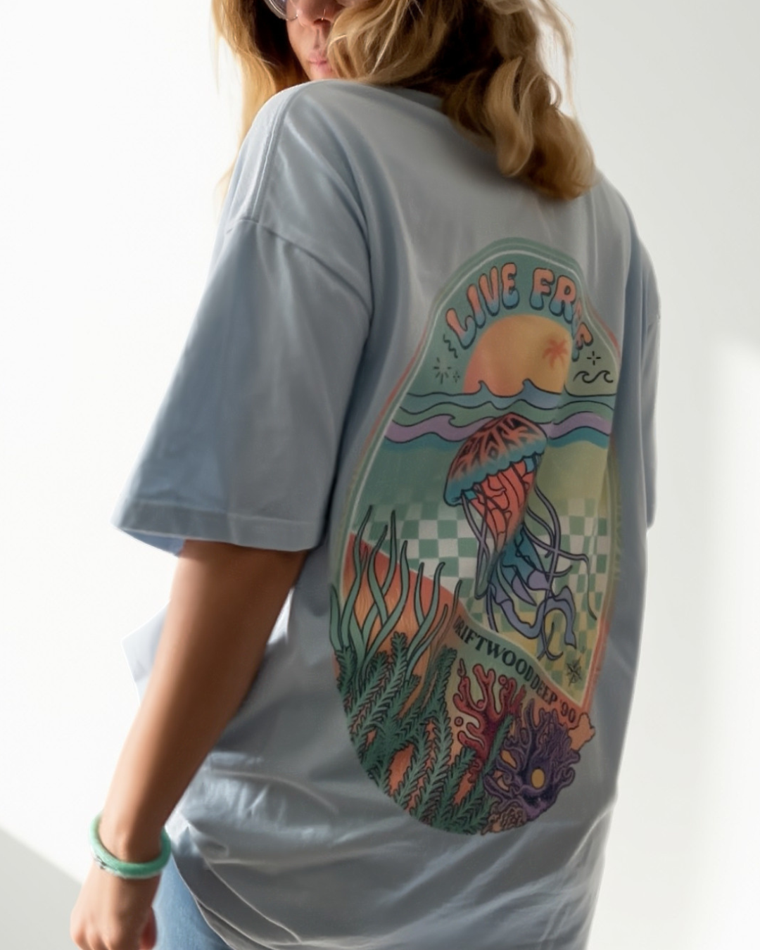 A scuba-diving woman wearing a light blue oversized T-shirt made for female scuba divers, viewed from the back. The shirt features a large, colourful graphic print of a jellyfish in an underwater scene with the text 'LIVE FREE'.