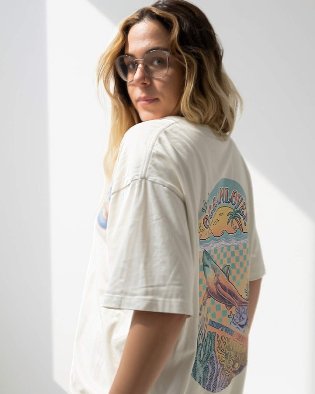 A woman scuba diver with blonde-highlighted hair and glasses, wearing an oversized off-white graphic T-shirt made for female scuba divers with a colourful shark illustration on the back, photographed in a bright studio setting.
