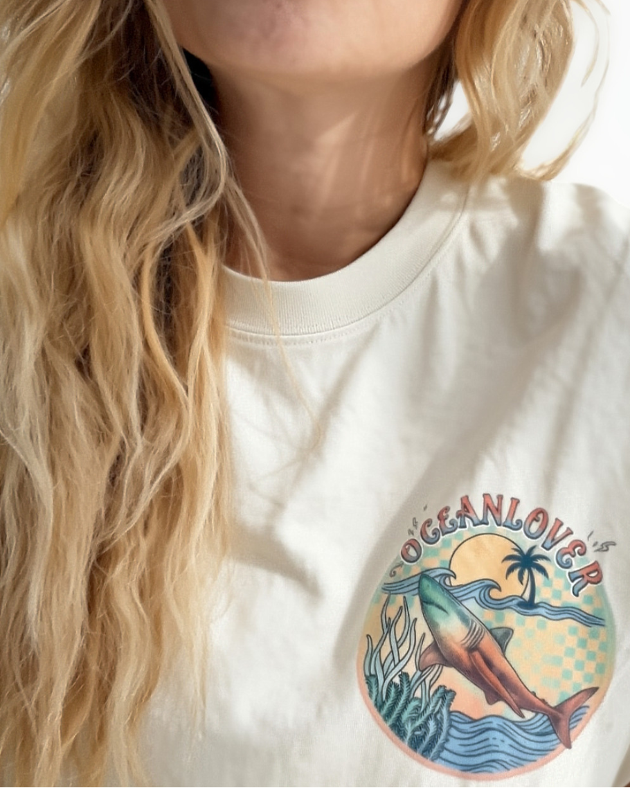 A close-up of a woman scuba diver wearing a cream-coloured T-shirt for female scuba divers with a colourful graphic on the left chest. The graphic features a shark, a palm tree, waves, and the text 'OCEANLOVER'.
