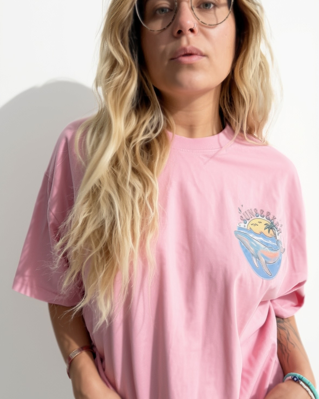 A woman with long blonde hair and glasses wearing a pink oversized T-shirt made for female scuba divers with a graphic on the left chest, set against a plain white wall.