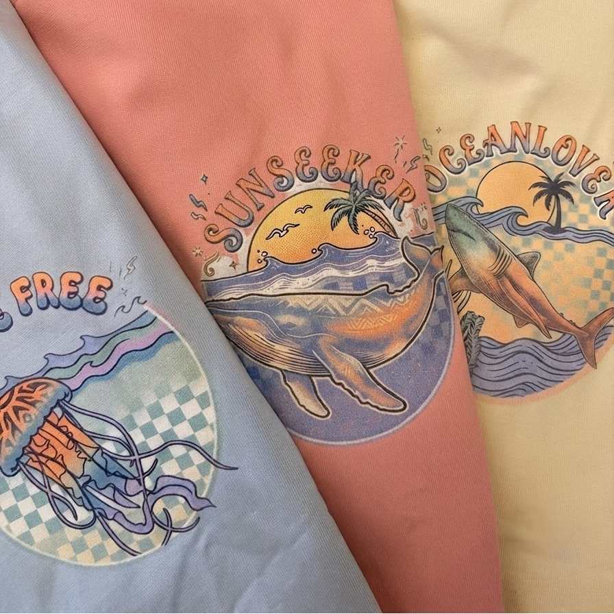 Three short-sleeve crewneck t-shirts in pastel blue, coral pink, and cream yellow, arranged diagonally. Each shirt features a colourful graphic print of a whale, dolphin, and jellyfish. 