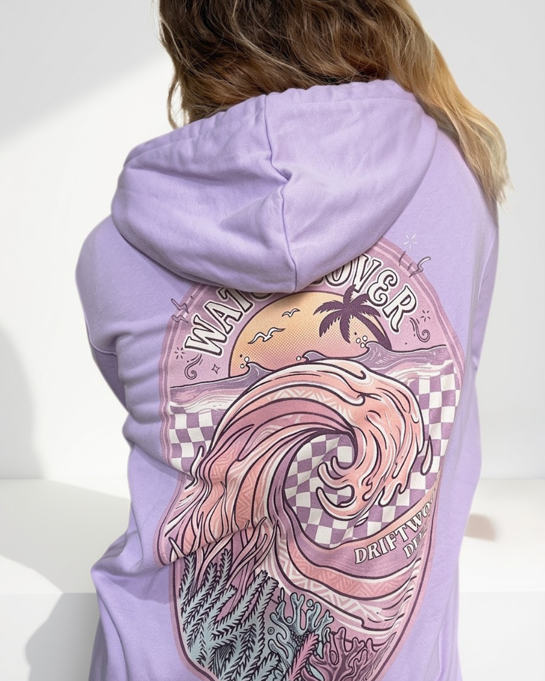A person shown from the back wearing a lavender hoodie with a large pink and peach graphic print featuring a wave, palm tree, and text on the back.