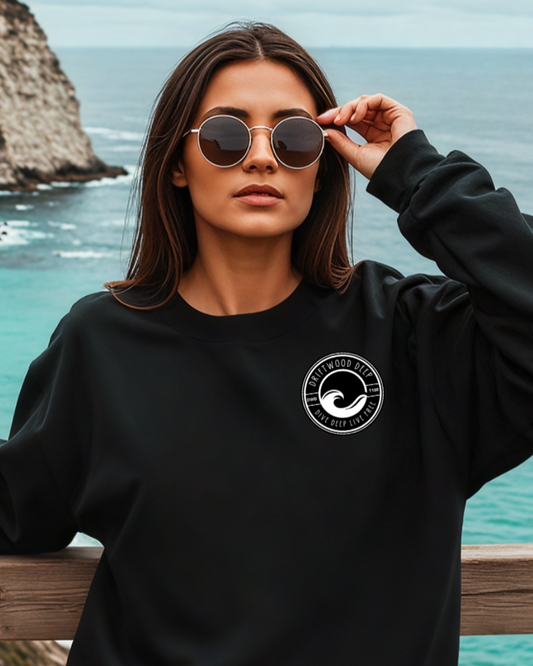 Woman wearing a black sweatshirt with a logo, standing by a coastal view.
