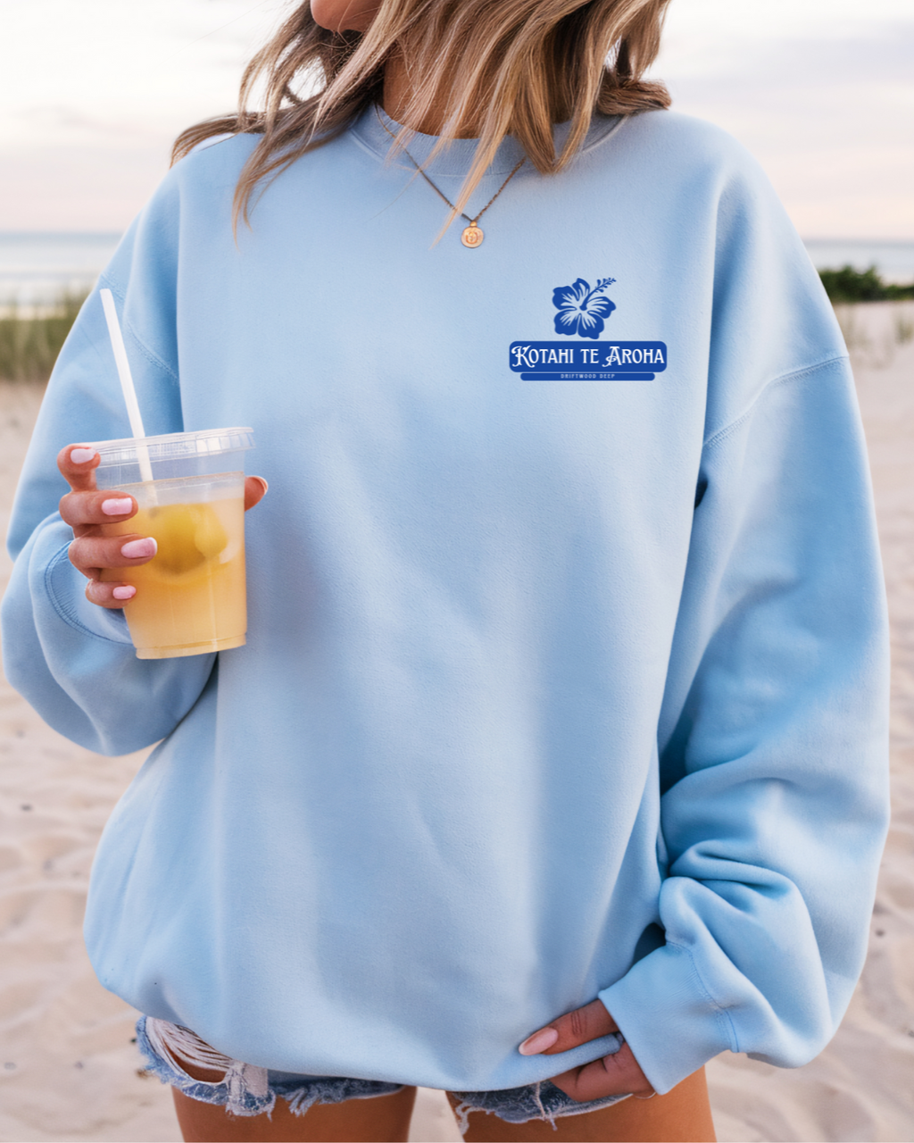 Person wearing a light blue sweatshirt with a logo, holding a drink outdoors.