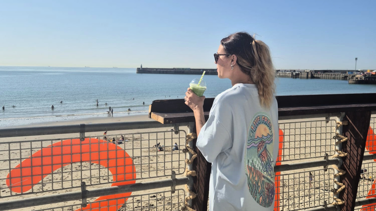 A woman stands on a boardwalk by the beach wearing a very oversized blue scuba diving graphic T-shirt with a colourful jellyfish print on the back, holding a drink. The background shows the ocean, sand, and a large orange graphic on the railing.
