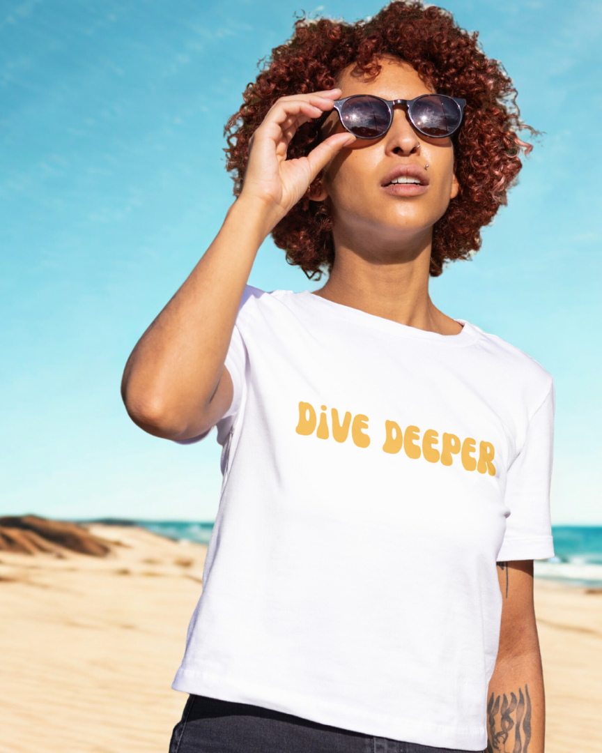 Woman on a beach wearing a white t-shirt with 'Dive Deeper' text, holding sunglasses.