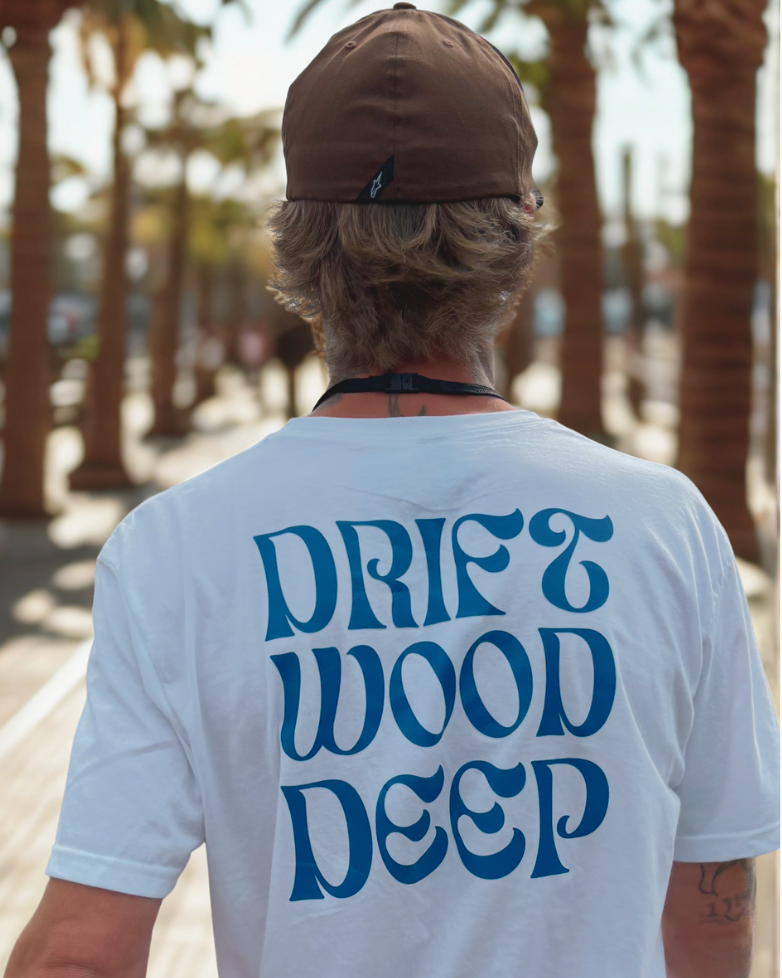 Person wearing a white t-shirt with 'Drift Wood Deep' text, standing in a palm tree-lined area.
