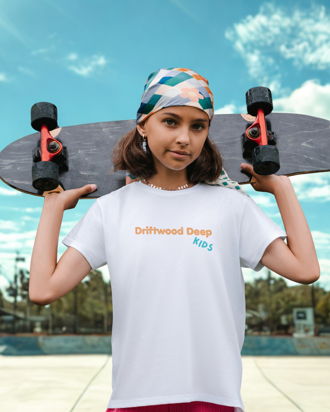 Child holding a skateboard with 'Driftwood Deep Kids' t-shirt against a clear sky.
