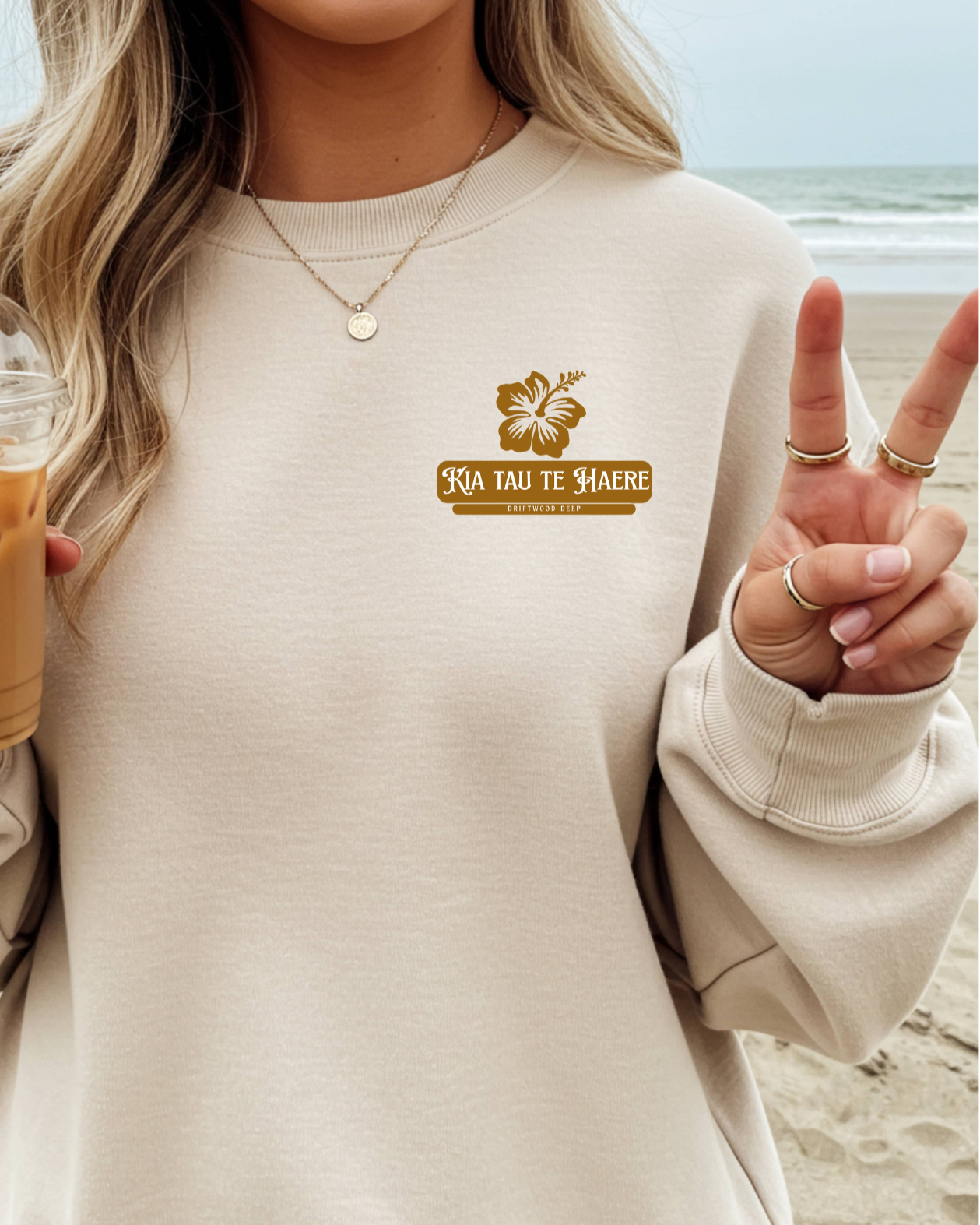 Person wearing a beige sweatshirt with a logo and text, holding an iced coffee on a beach.