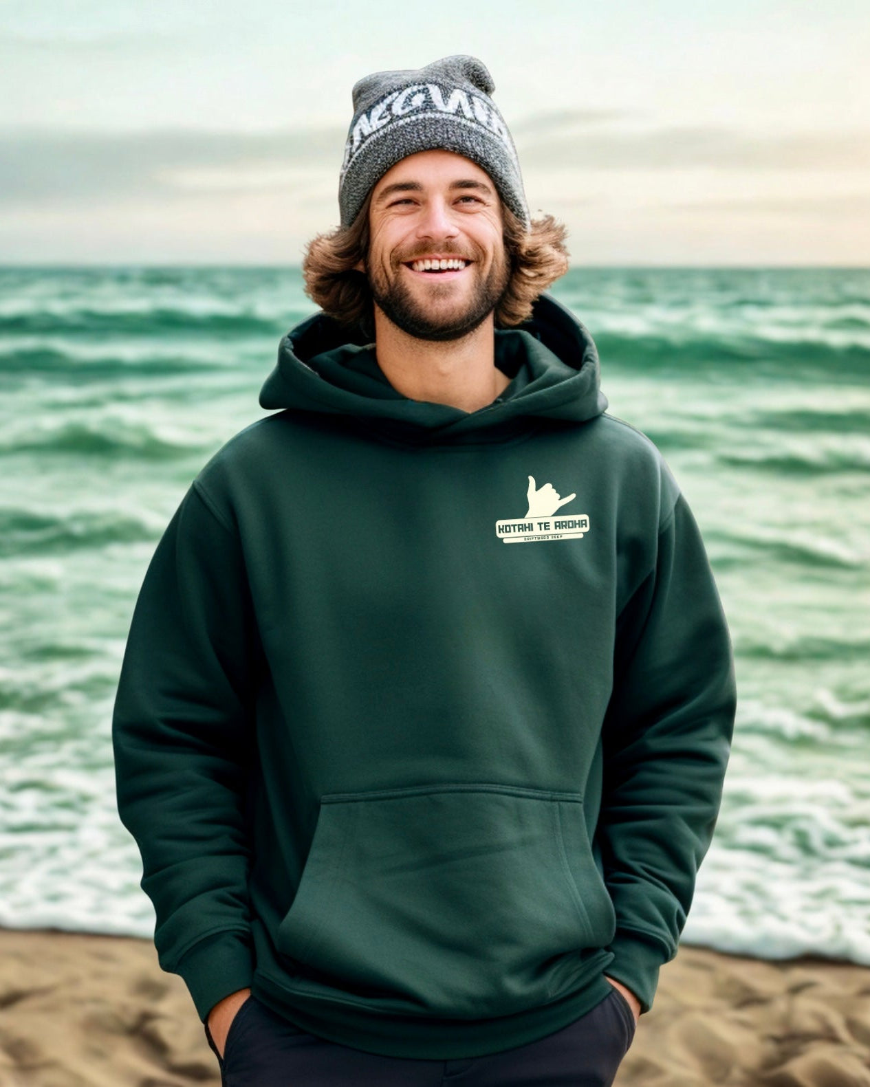 Man wearing a green hoodie and beanie standing on a beach with ocean waves in the background