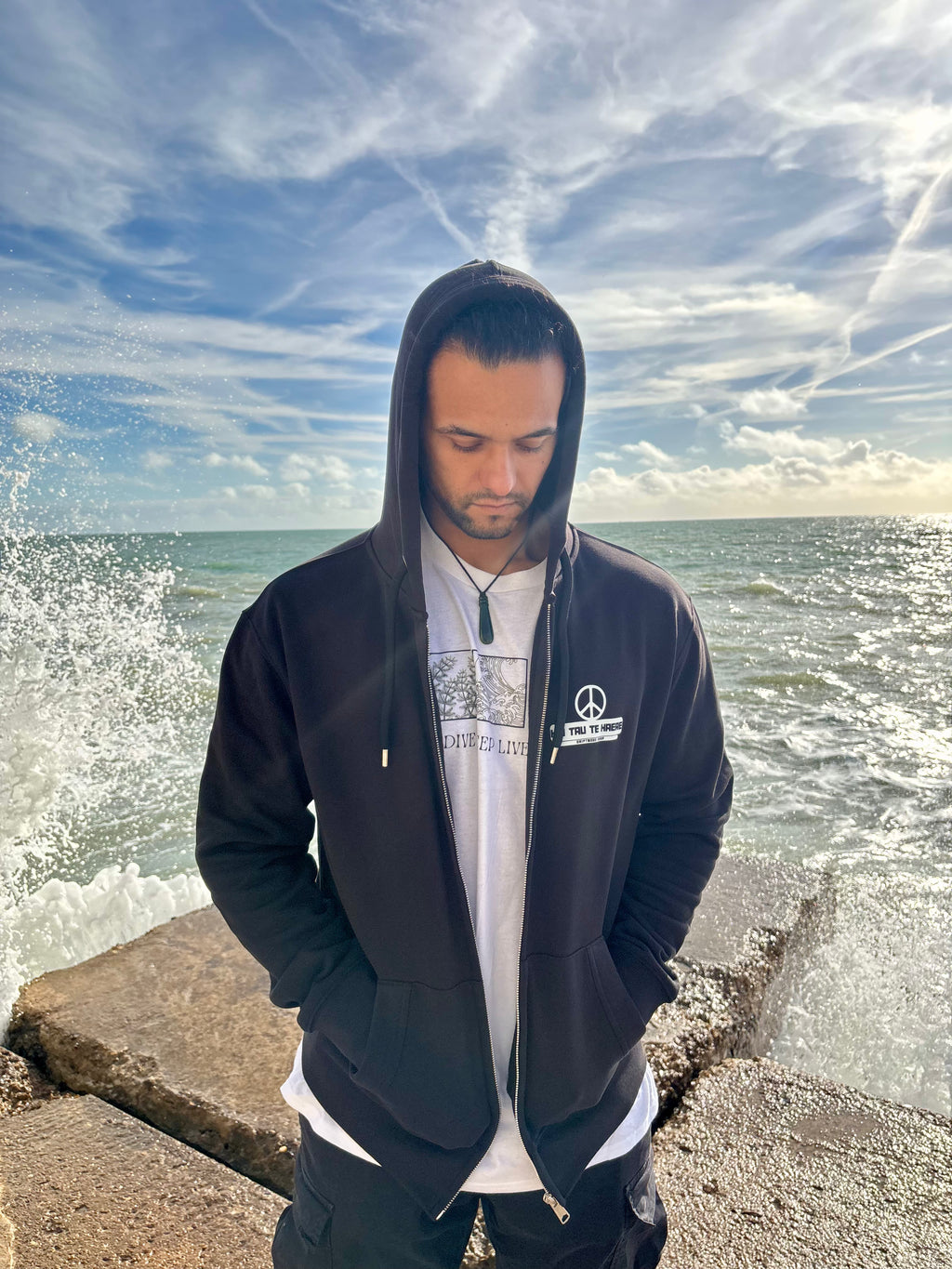 Man wearing a dark hoodie and white shirt with a logo, standing by the ocean.