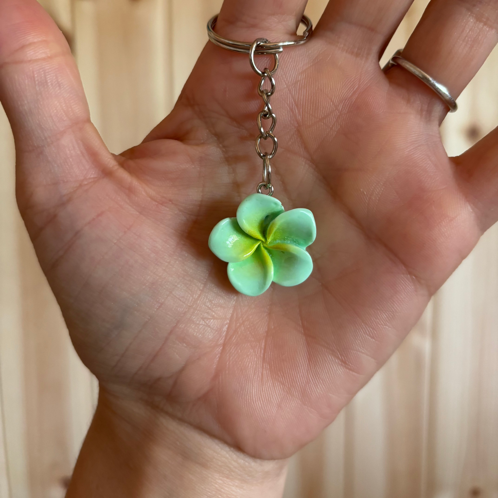 Hand holding a keychain with a green flower charm against a neutral background