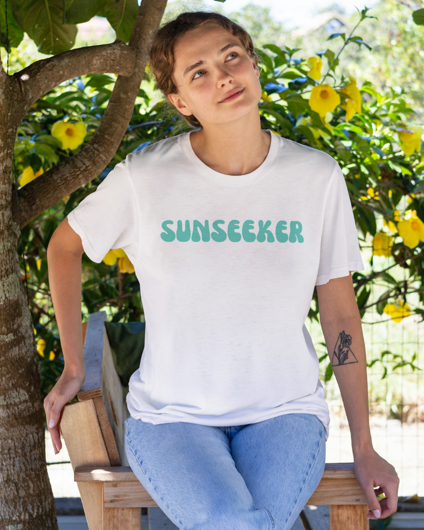 Person wearing a white t-shirt with 'Sunseeker' text, sitting under a tree with flowers.
