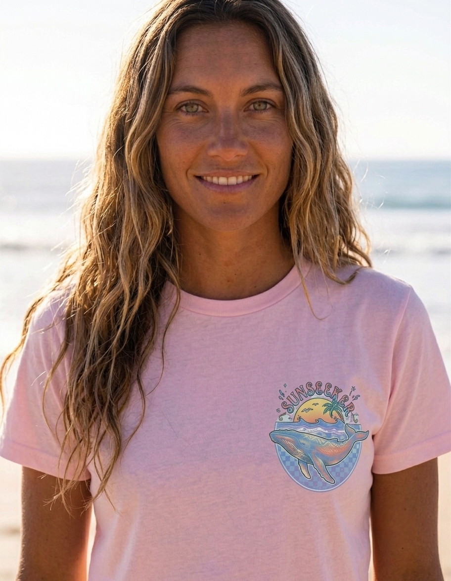 A woman with long wavy light brown hair stands on a beach wearing a light pink t-shirt with a whale graphic on the left chest. The graphic features a blue whale under a yellow sun and palm tree, with sunseeker written in arched yellow text.