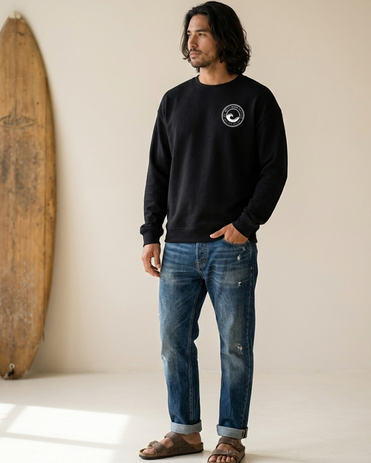 Man wearing a black sweatshirt with a logo and blue jeans standing next to a wooden surfboard against a white wall.