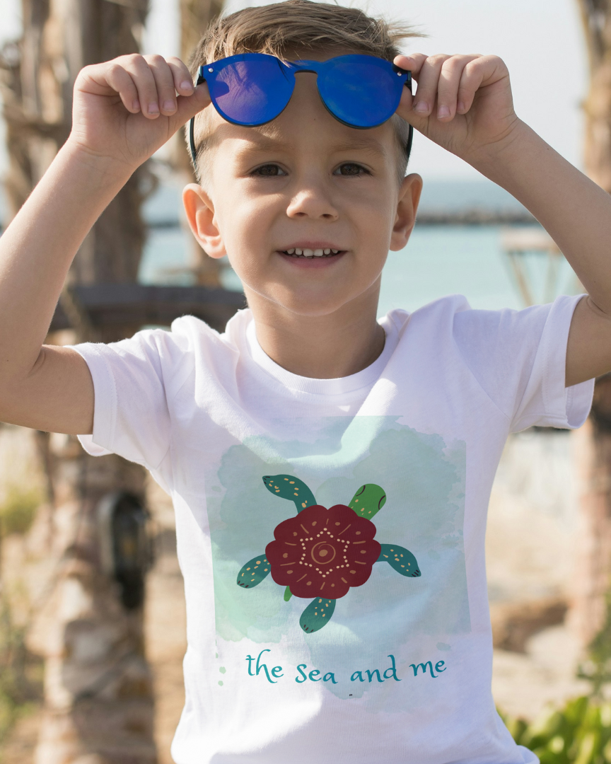 Child wearing a white t-shirt with a turtle design and text, holding blue sunglasses.