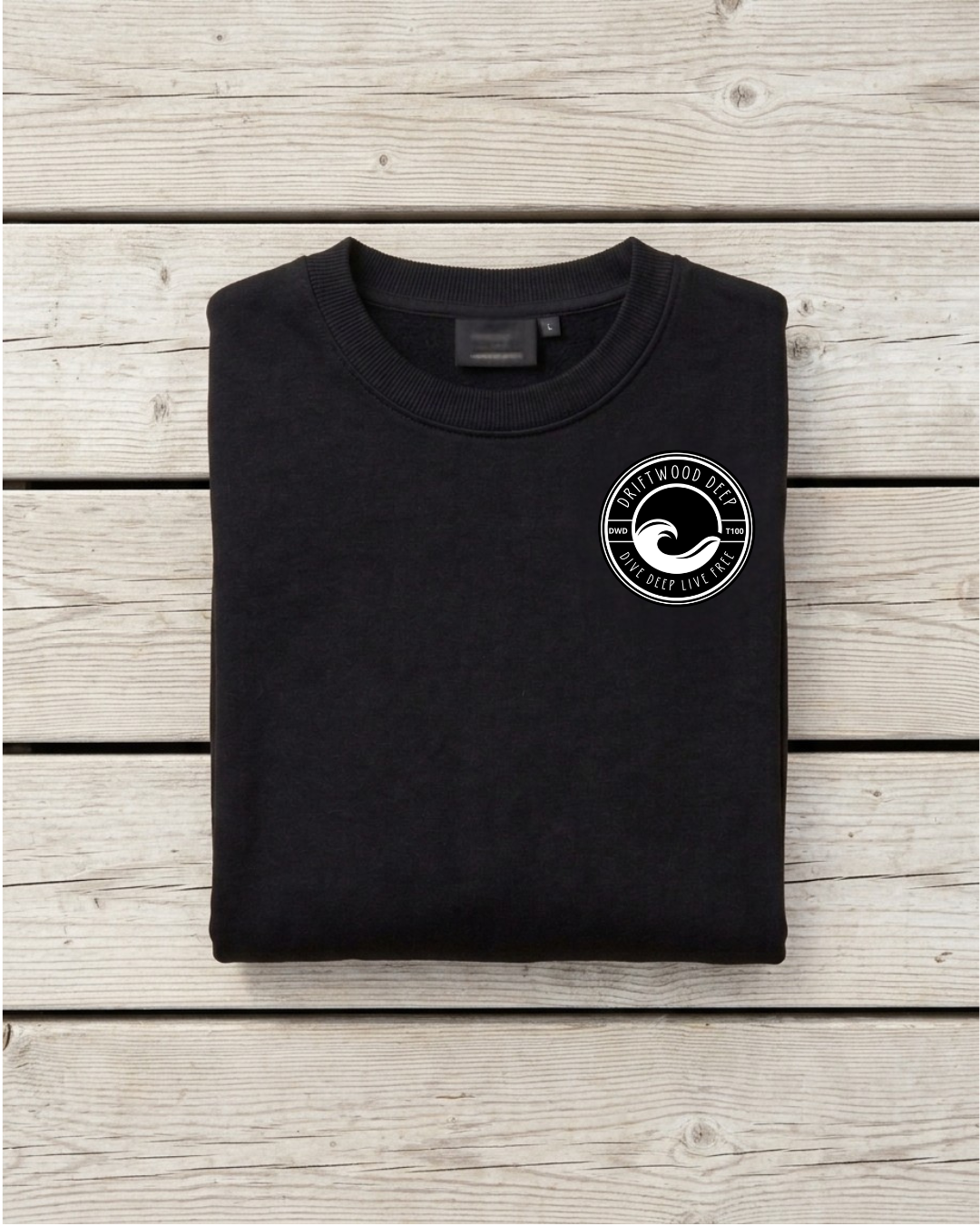 A folded black crewneck t-shirt with a white circular wave logo on the left chest, displayed flat on a light gray wooden plank background.