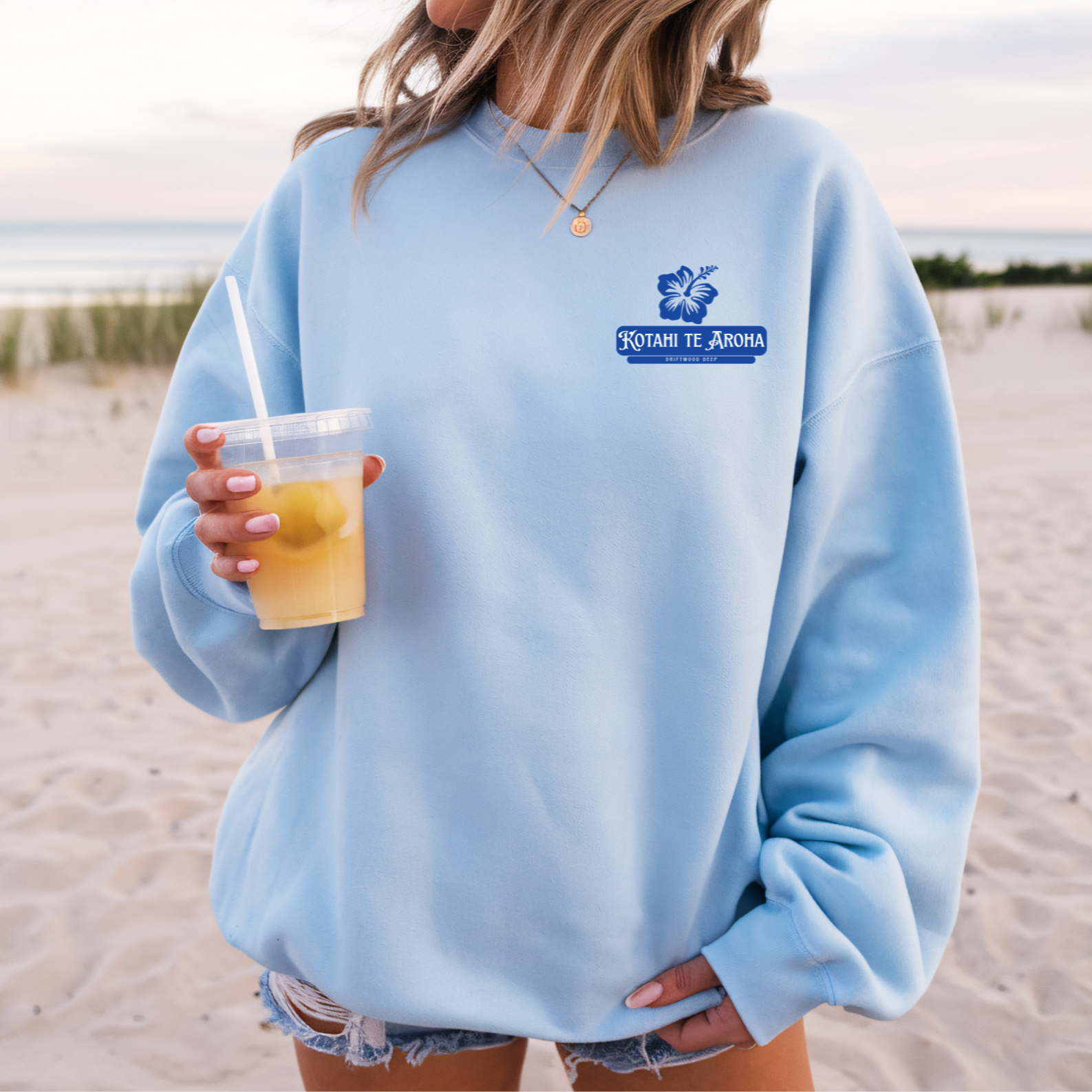 Person wearing a light blue sweatshirt with a logo, holding a drink outdoors.