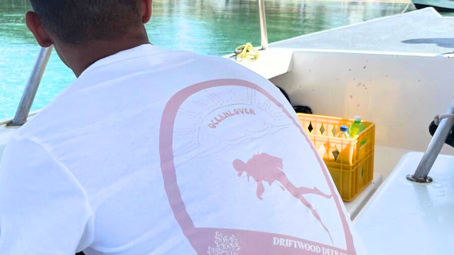 Person on a boat wearing a white shirt with a pink graphic design, sitting next to a yellow crate.