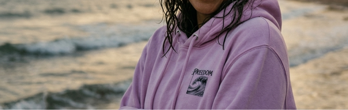 A smiling woman with wet dark hair stands on a beach wearing a light purple hoodie with a black 'FREEDOM' wave logo on the left chest. The background shows ocean waves and a rocky shoreline at sunset.