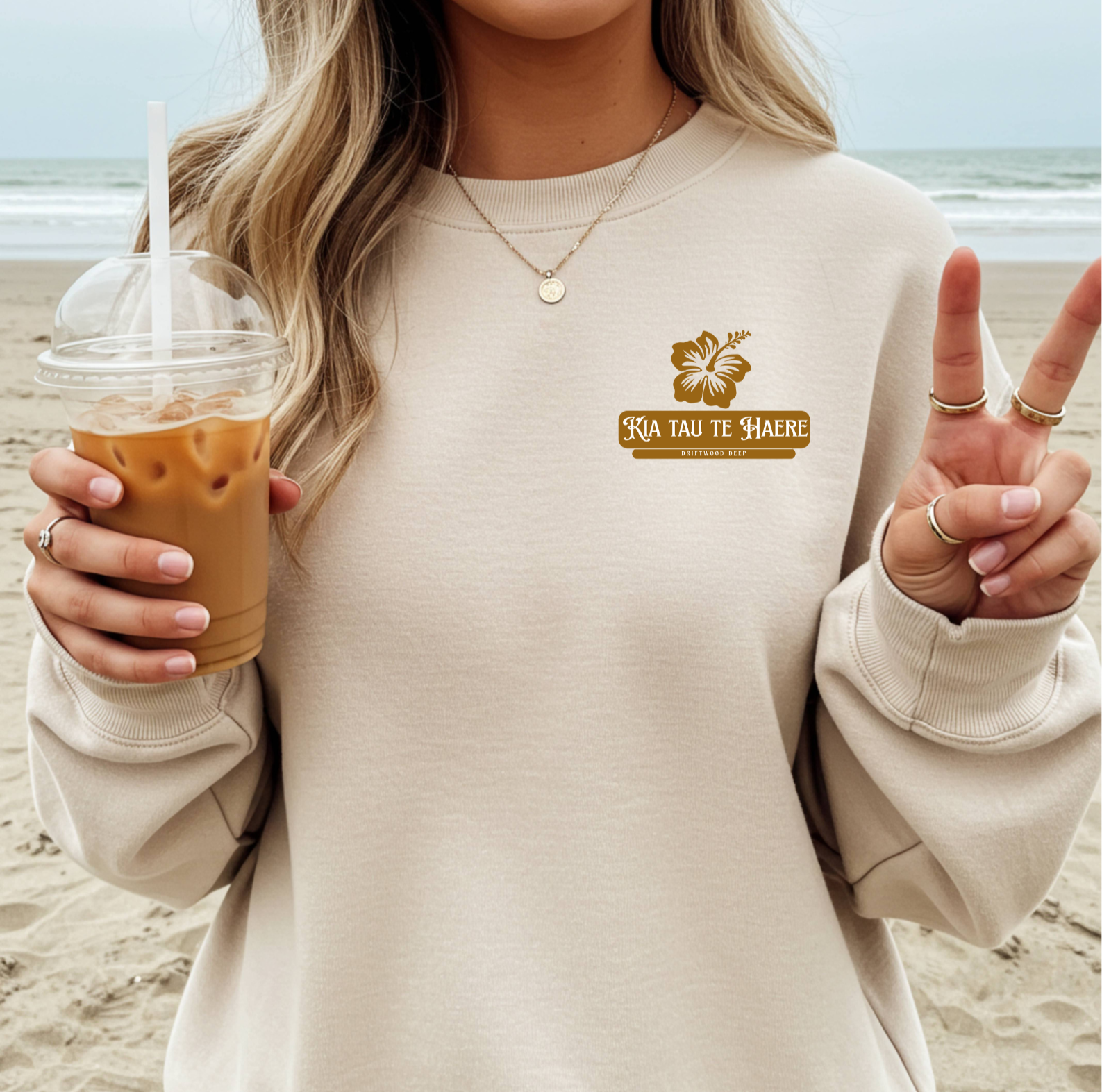 Person wearing a beige sweatshirt with a logo and text, holding an iced coffee on a beach.